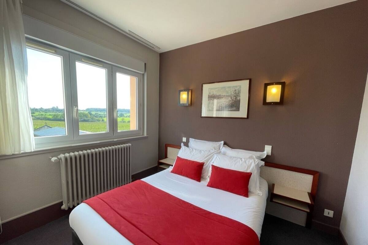 Habitación de hotel con una cama grande, vistas al campo a través de una ventana, un radiador y dos luces fijadas a la pared.