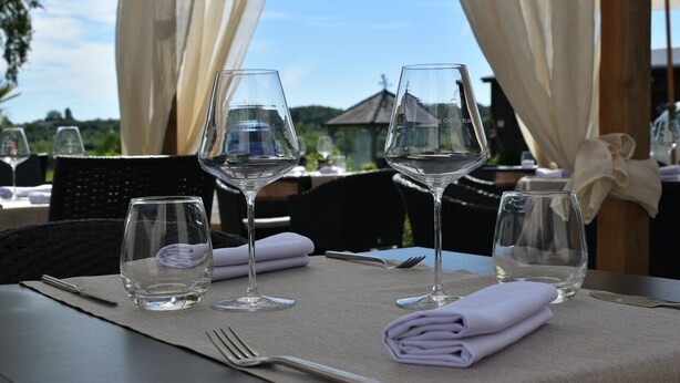 Une table dressée pour le dîner, avec des verres à vin et des couverts, recouverte de rideaux blancs et offrant une vue sur la verdure.