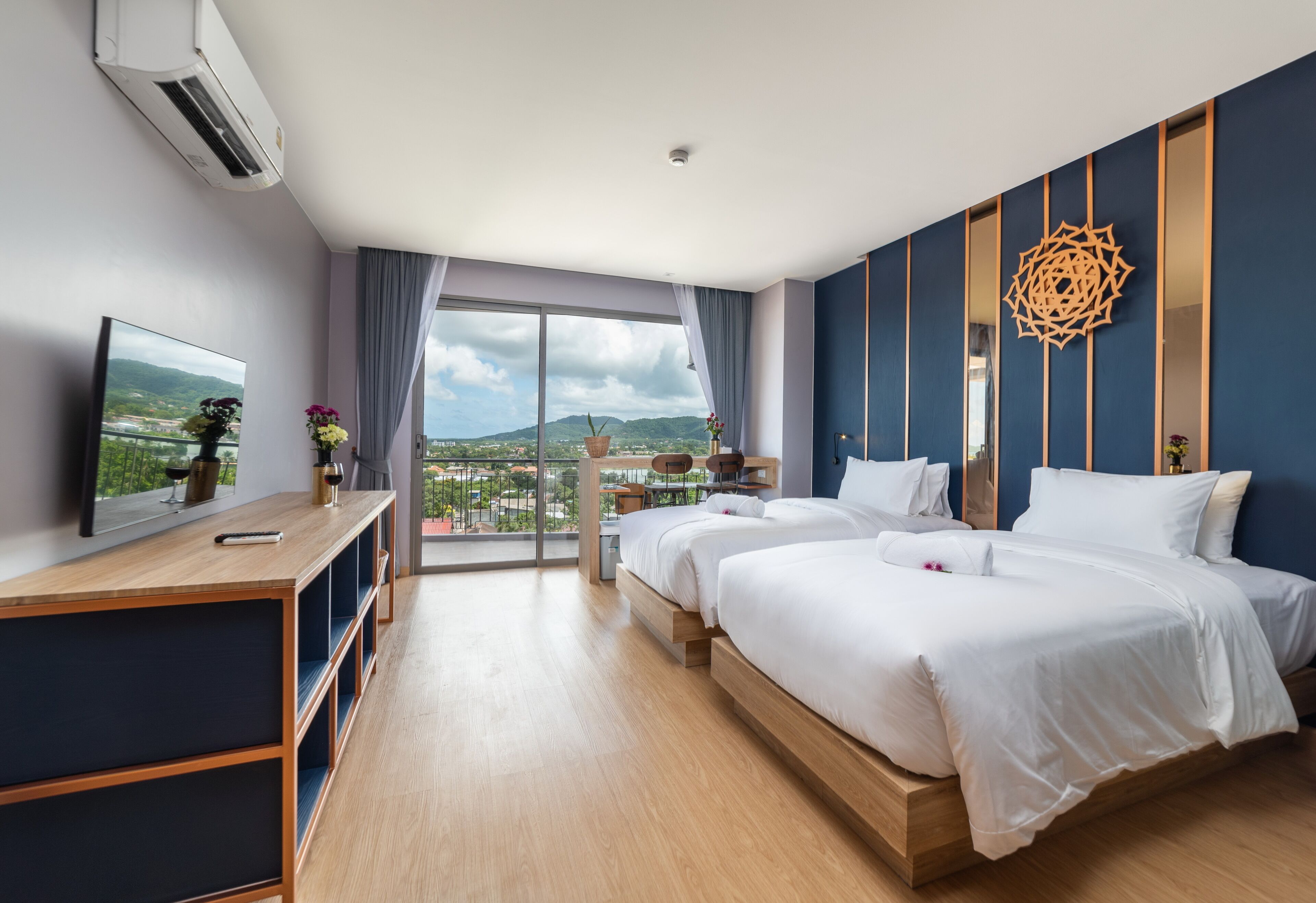 A modern hotel room with a large bed, a desk, and a balcony with a view of the city and mountains.