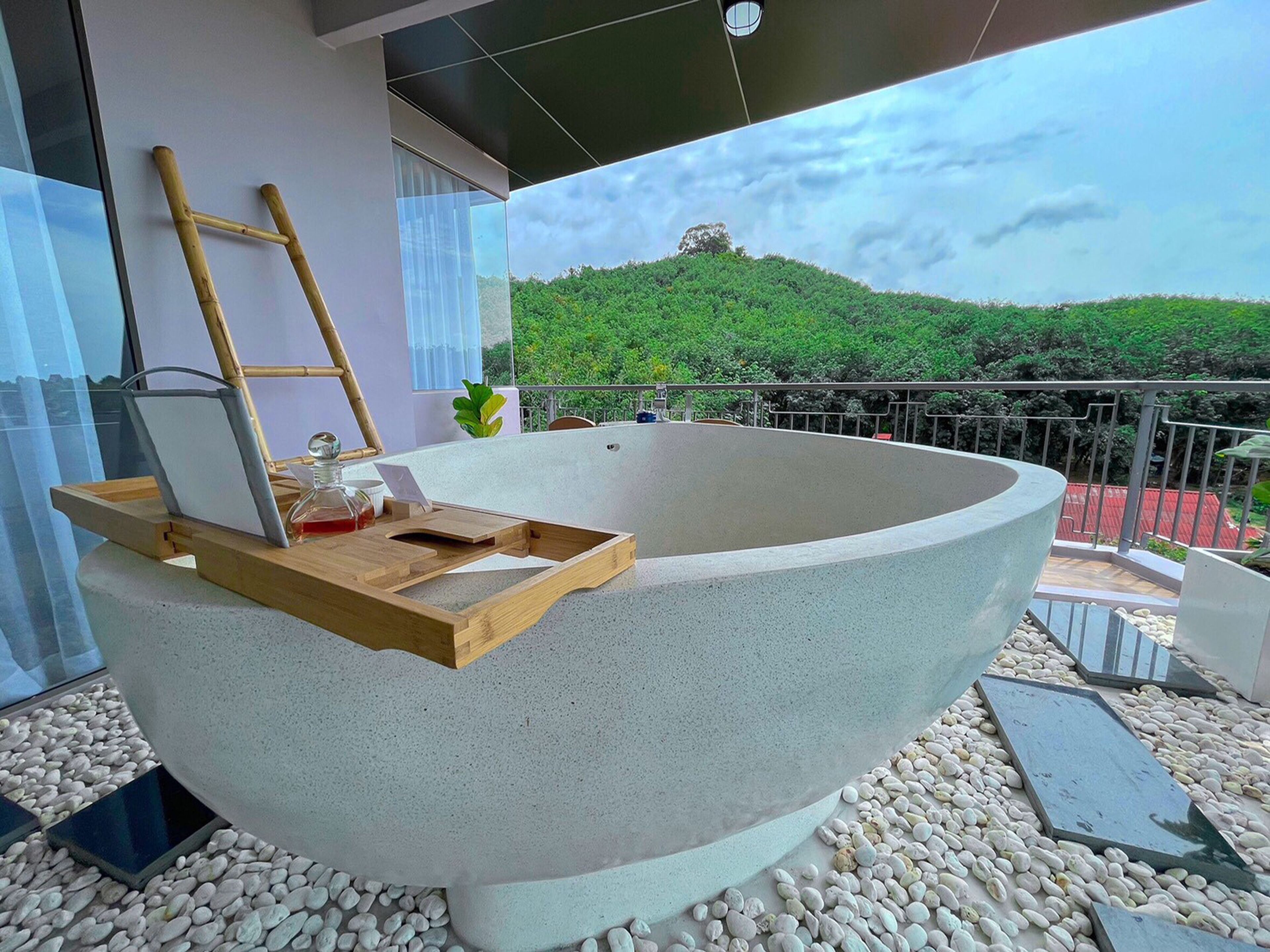 A modern outdoor bathtub with a wooden tray, a ladder, and a bottle on a balcony with a view of a green hill.
