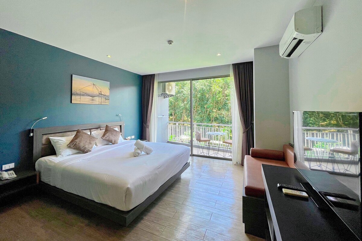 A hotel room with a bed, a desk, a chair, a balcony with a view of greenery, and an air conditioning unit.