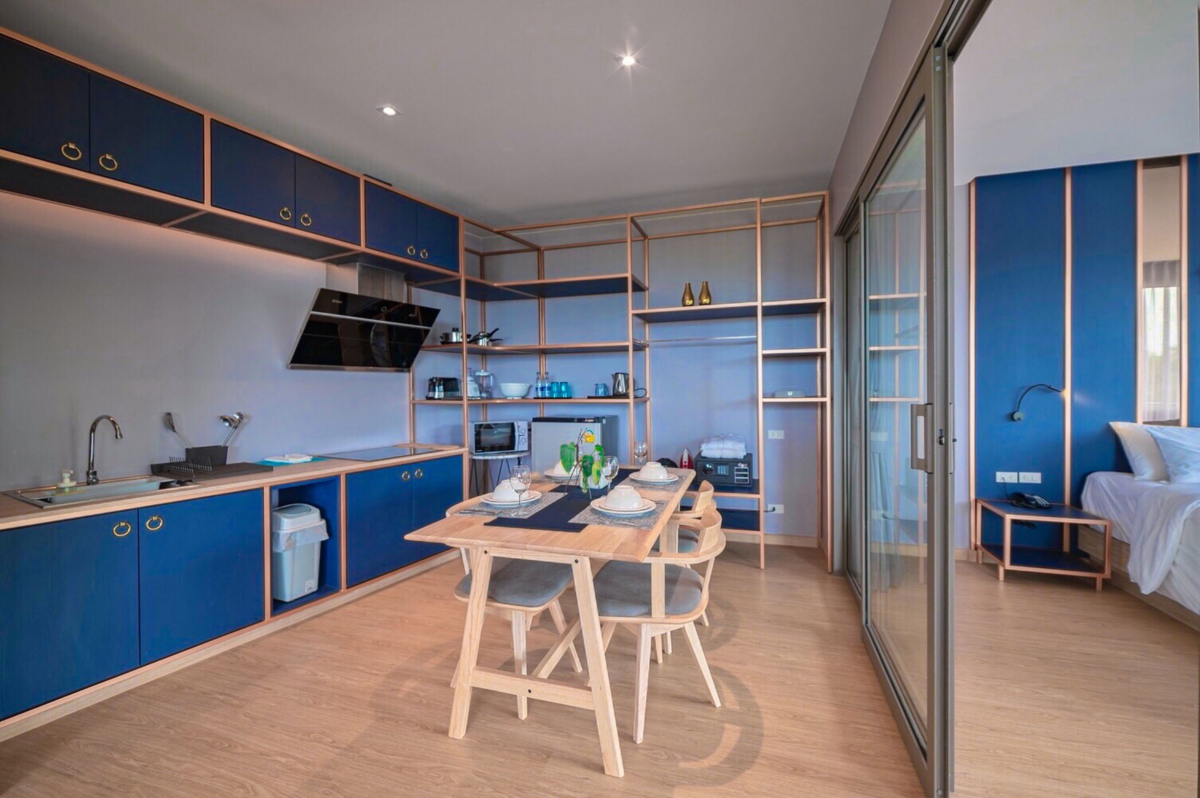 A modern kitchen with blue cabinets, a dining table set for four, and a glass-enclosed sleeping area with a bed and bedside table.