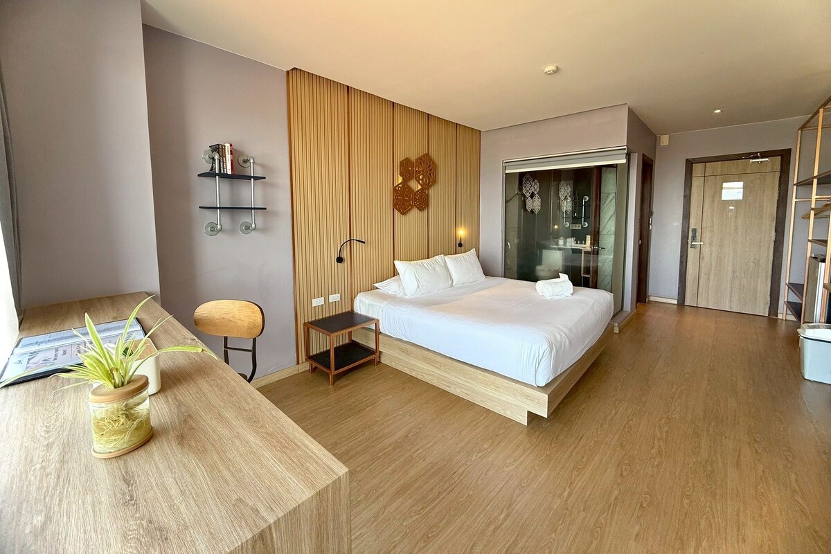A modern hotel room with a large bed, a desk with a plant, a small table, and a sliding door leading to a bathroom.