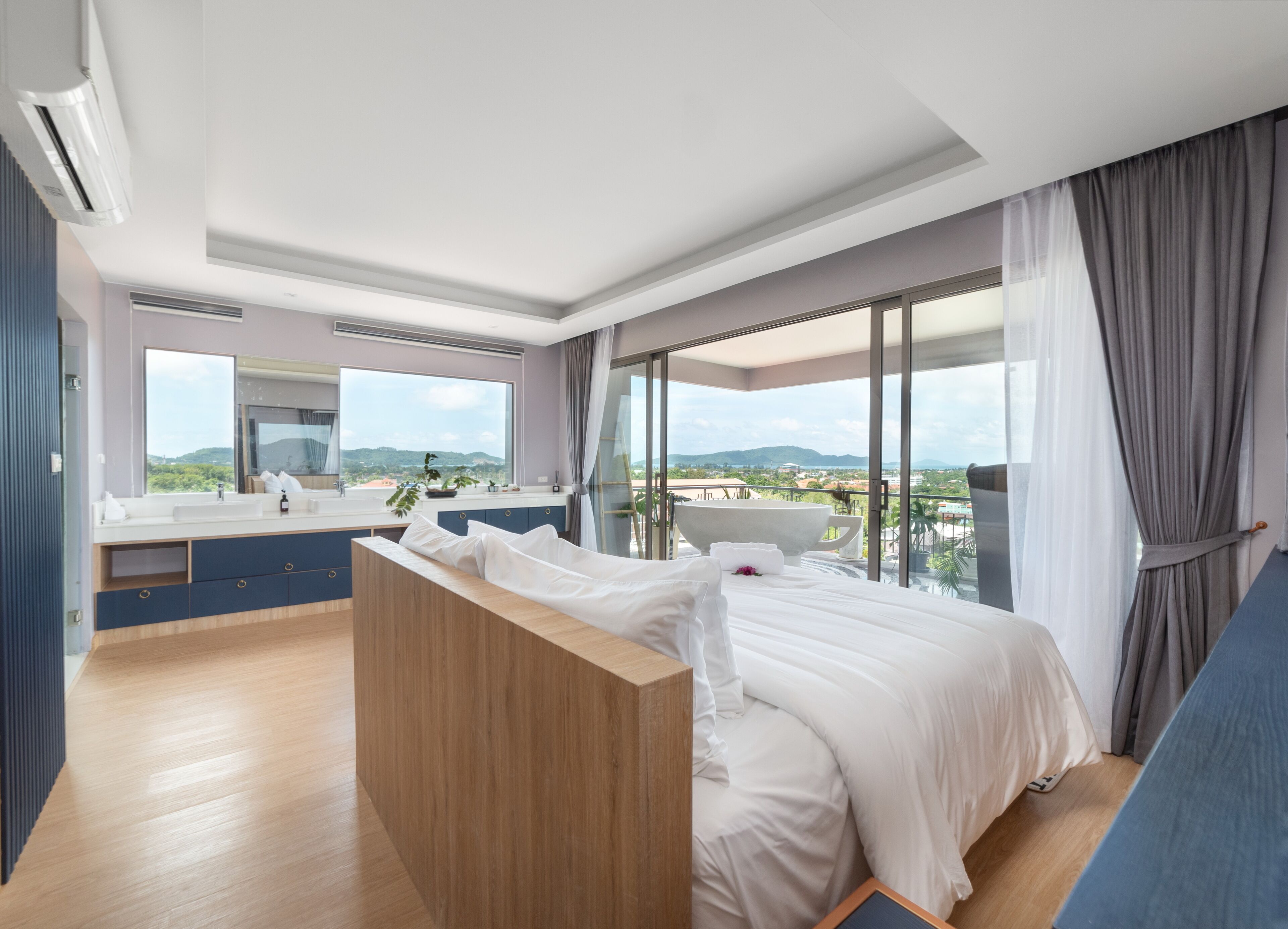 A modern hotel room with a large bed, a sitting area, a bathroom with a large mirror, and a view of the mountains through large windows.