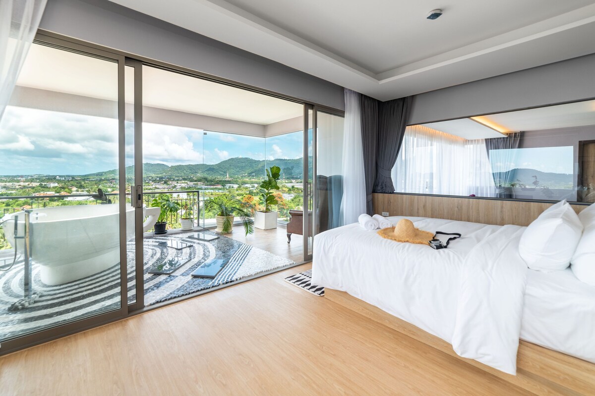 A modern hotel room with a large bed, a bathroom with a bathtub, and a balcony with a view of the city and mountains.