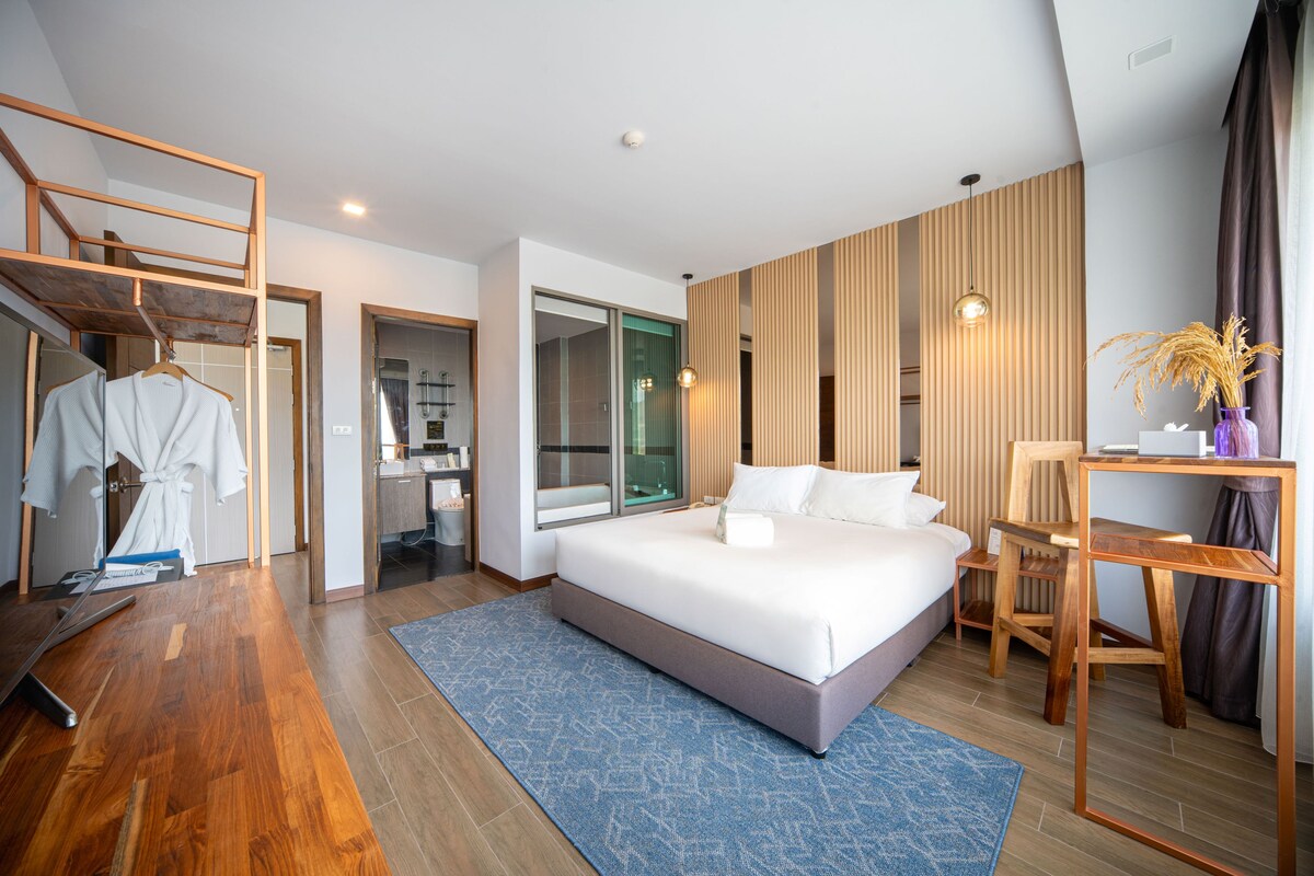 A modern hotel room with a large bed, a wooden desk, a wardrobe, and a bathroom visible through an open door.