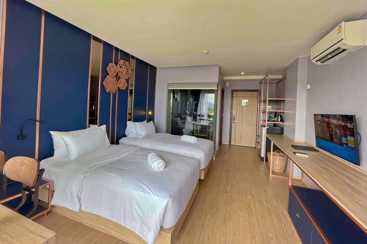 A hotel room with two beds, a desk, a chair, a TV, and an air conditioning unit.