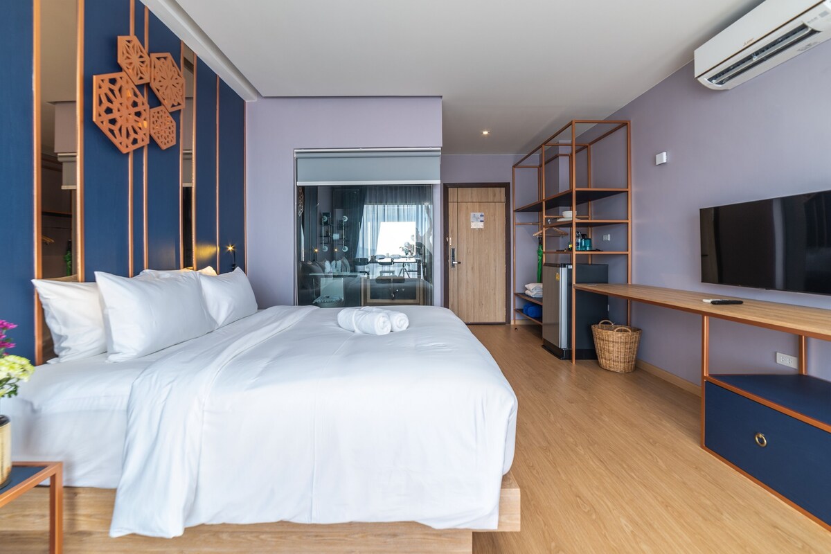 A modern hotel room with a large bed, a TV, a wooden bookshelf, and a glass door leading to a dining area.