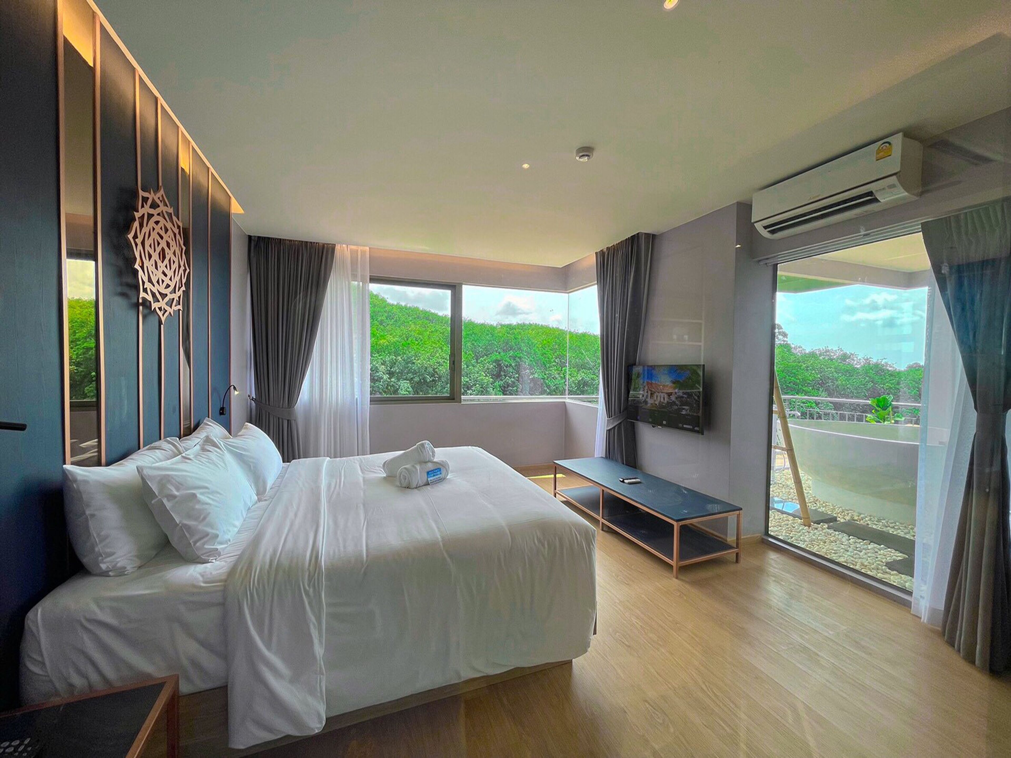 A modern hotel room with a large bed, a TV, and a balcony with a view of greenery.