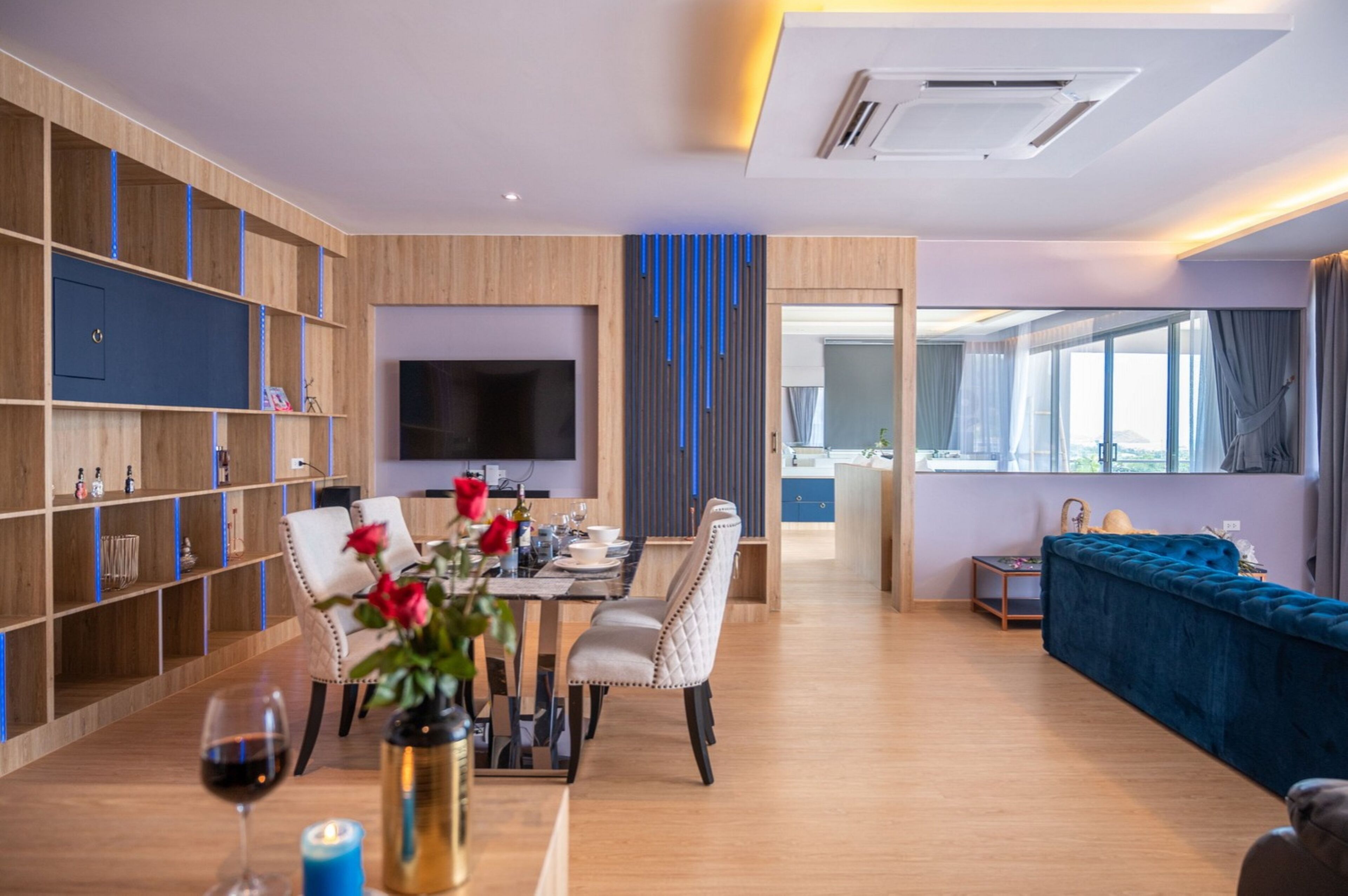 A modern living room with a dining area, wooden shelving, a flat-screen TV, and a blue sofa.