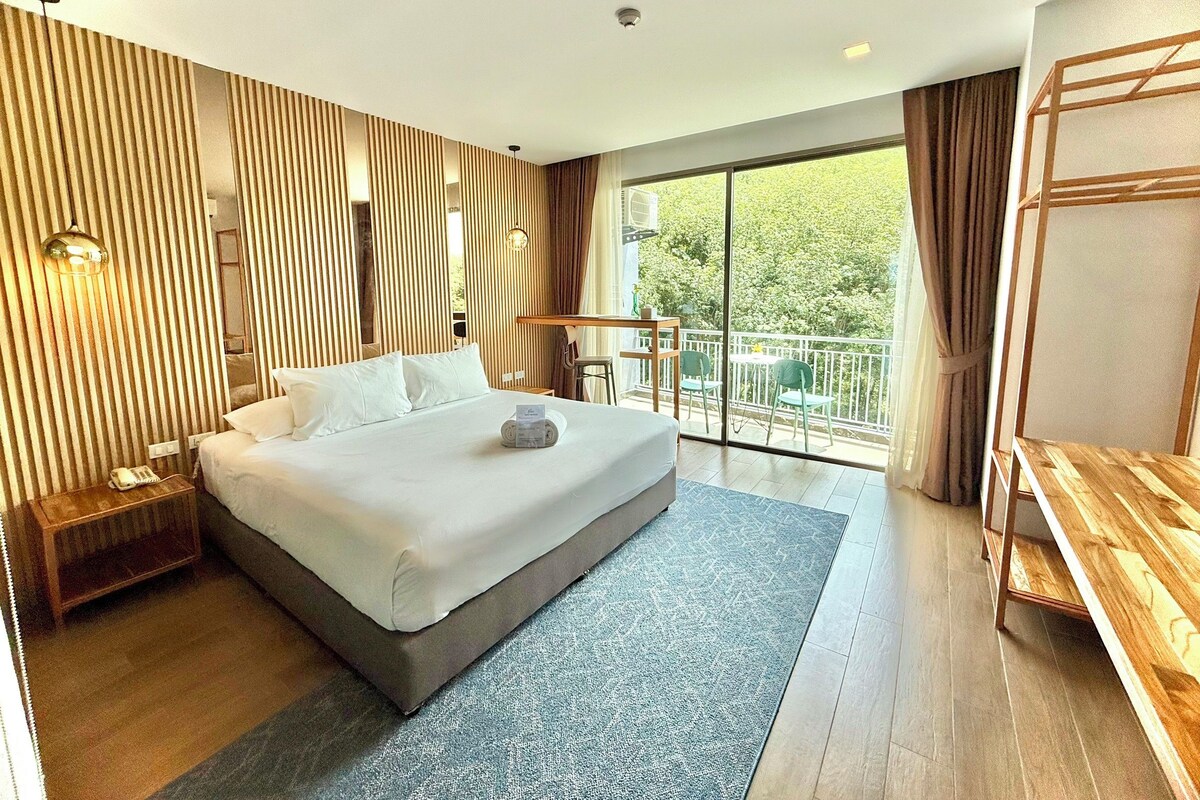 A modern hotel room with a large bed, a balcony with a table and chairs, and a wooden desk.