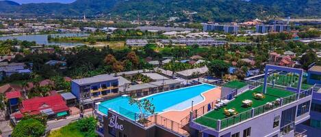A hotel with a rooftop pool, surrounded by greenery and a view of a lake and mountains.