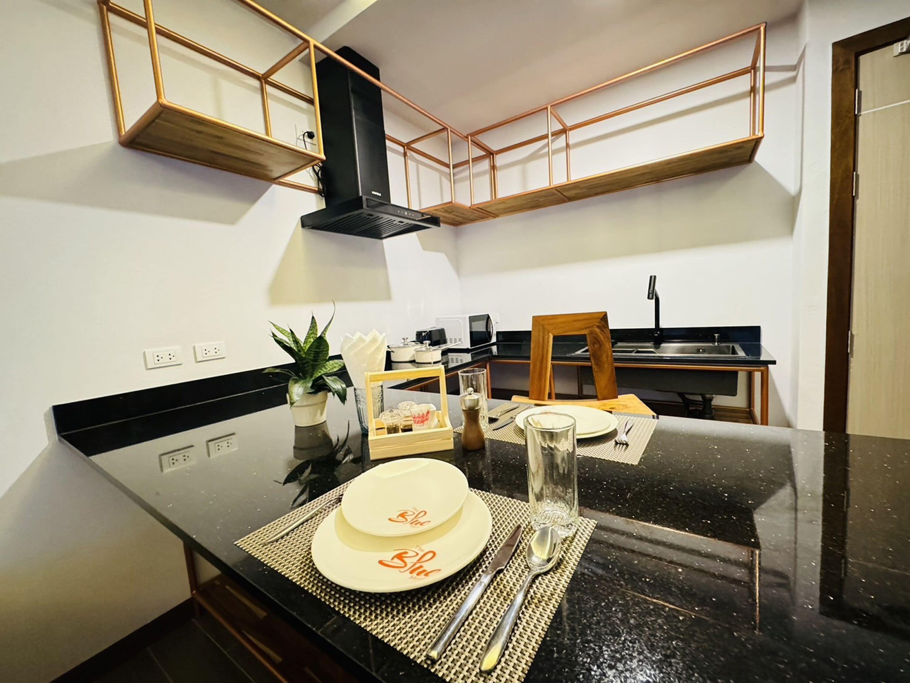 A modern kitchen with black countertops, wooden cabinets, and a black range hood. There is a dining table set with plates, cutlery, and a glass.