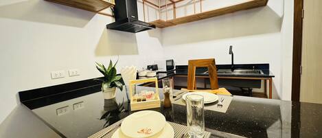 A modern kitchen with black countertops, wooden cabinets, and a black range hood. There is a dining table set with plates, cutlery, and a glass.