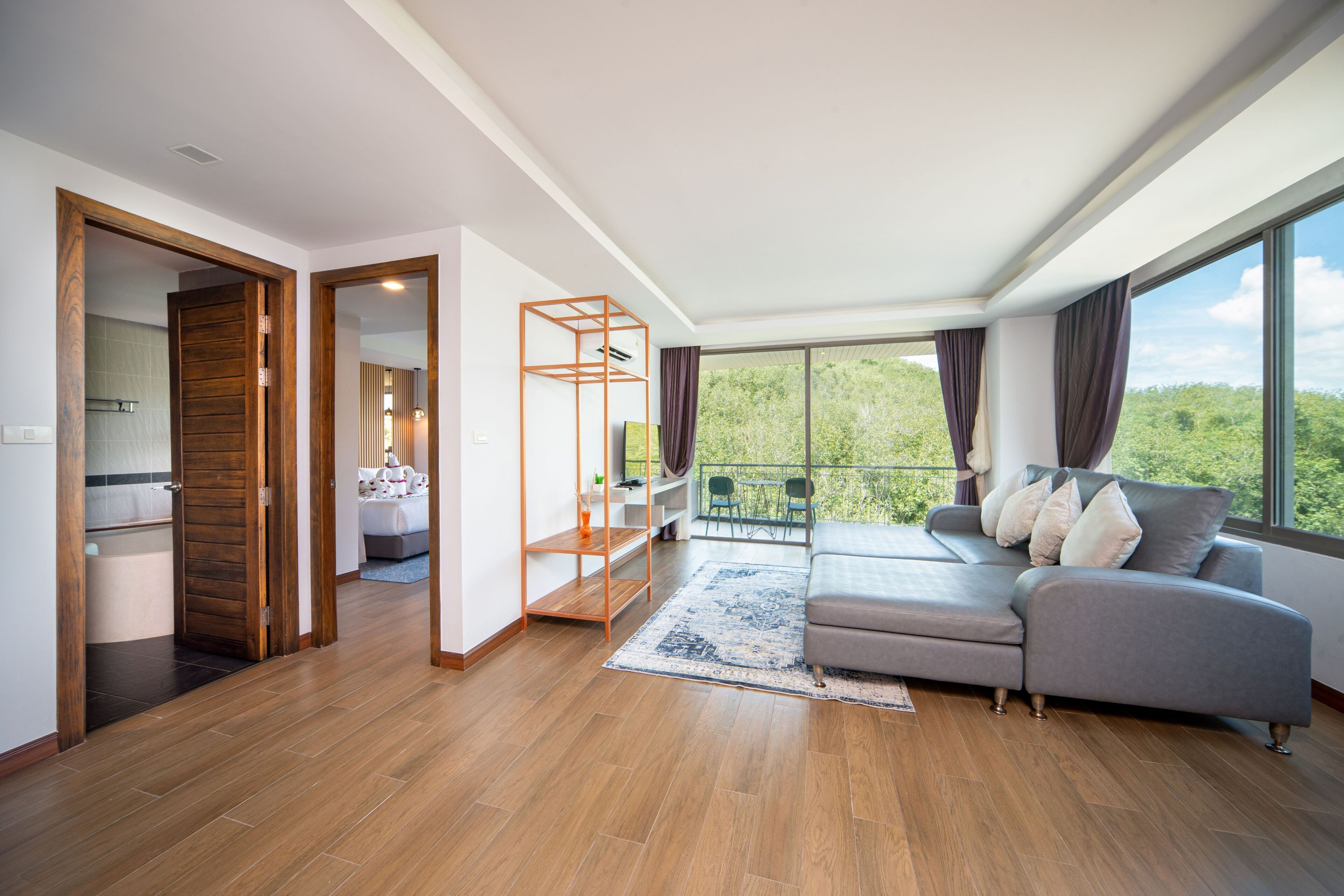 A spacious living room with a grey sofa, wooden flooring, and a large window offering a view of greenery.