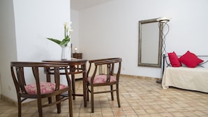 Un comedor con una mesa redonda y cuatro sillas de madera con cojines estampados. Un jarrón con flores blancas está sobre la mesa. En el fondo, una cama con colcha blanca y dos almohadas rojas.