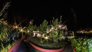 Vista nocturna de un área ajardinada con un sendero curvo, árboles y un edificio con ventanas iluminadas.