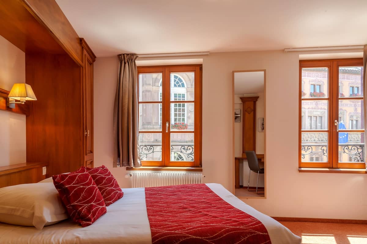 A hotel room with a large bed, a desk, and a view of the city through large windows.