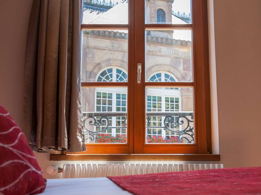 A hotel room with a large window, a radiator below, and a bed with a red blanket.