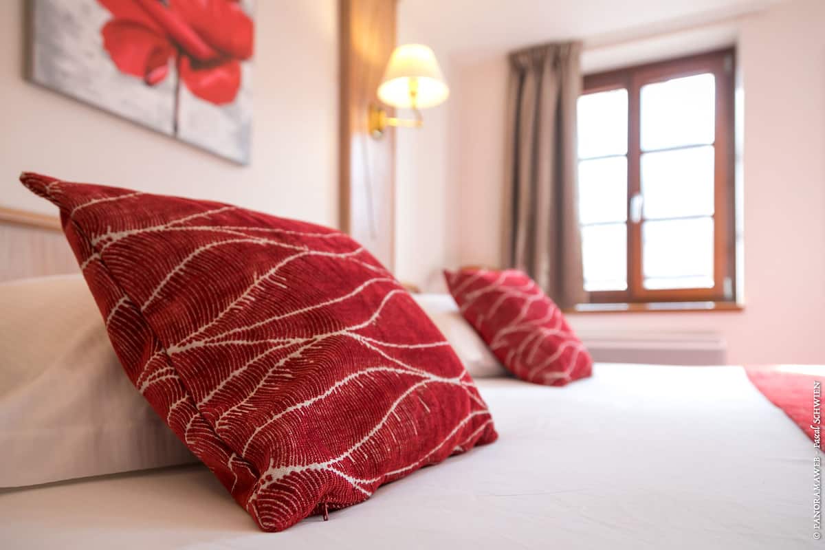 A hotel room with a bed, red patterned pillows, a painting on the wall, and a window with curtains.