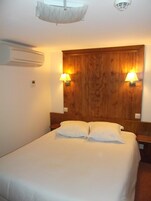 Habitación de hotel con cabecera de madera, dos lámparas de noche, cama con ropa blanca y un equipo de aire acondicionado en la pared.