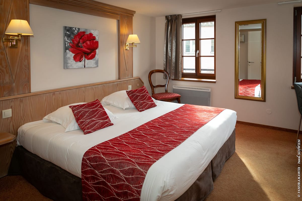 A hotel room with a large bed, two bedside lamps, a window with curtains, a wooden headboard, a red and white patterned bedspread, and a painting of red flowers on the wall.