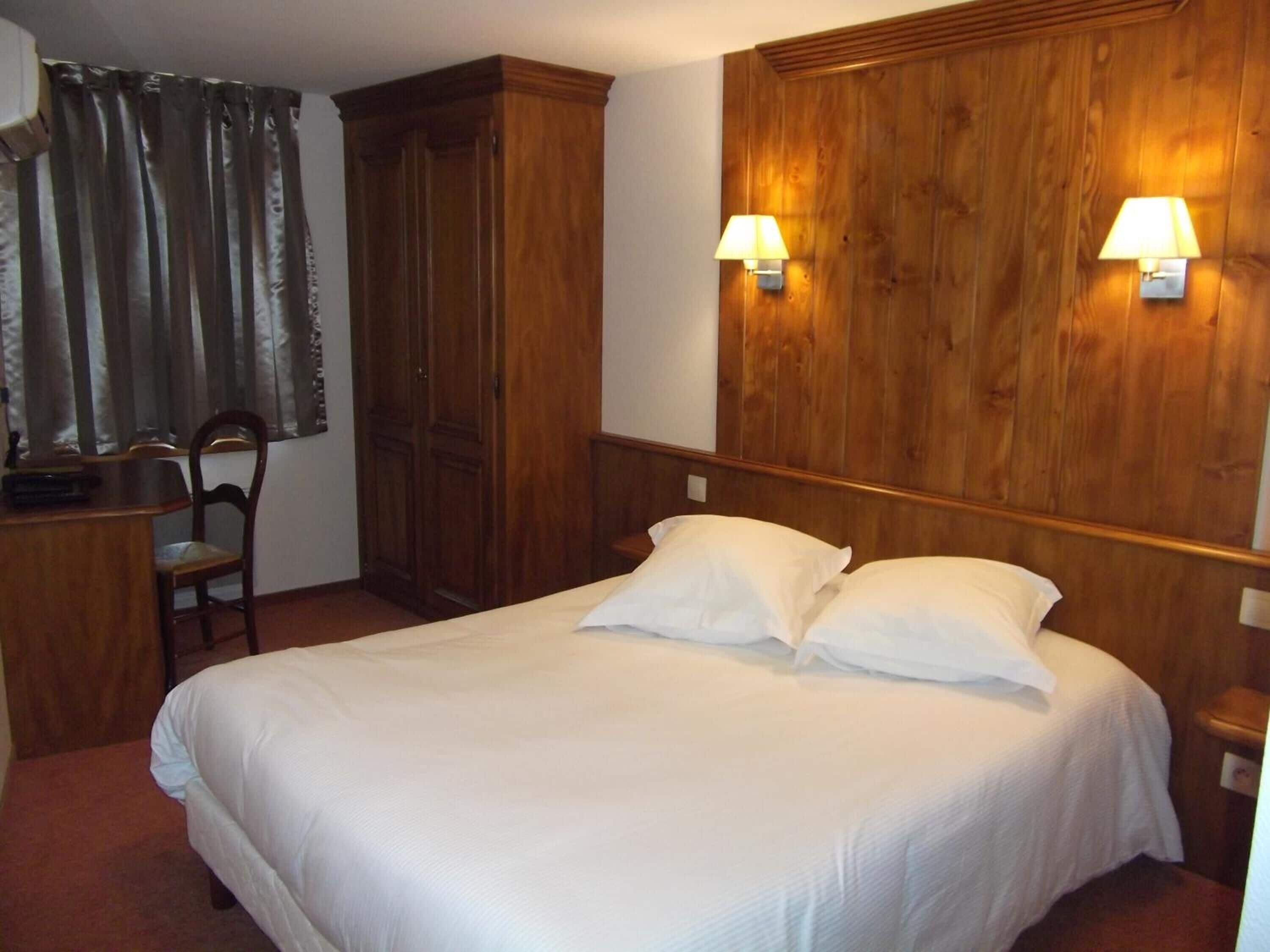 A hotel room with a large bed, wooden headboard, two bedside lamps, a wooden wardrobe, a desk with a chair, and a window with curtains.