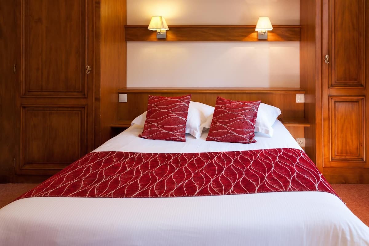 A hotel room with a large bed, two bedside lamps, wooden paneling, and a red and white patterned bedspread.