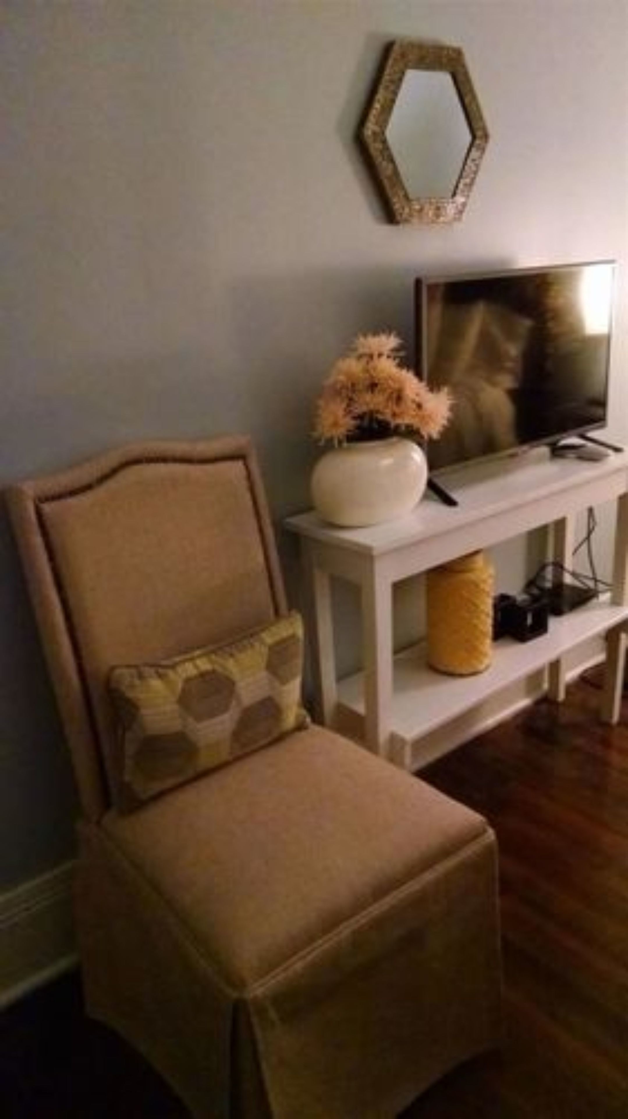 Una sala de estar con un sillón beige, una mesita auxiliar blanca con un jarrón de flores secas, un televisor sobre un mueble blanco y un espejo hexagonal en la pared.