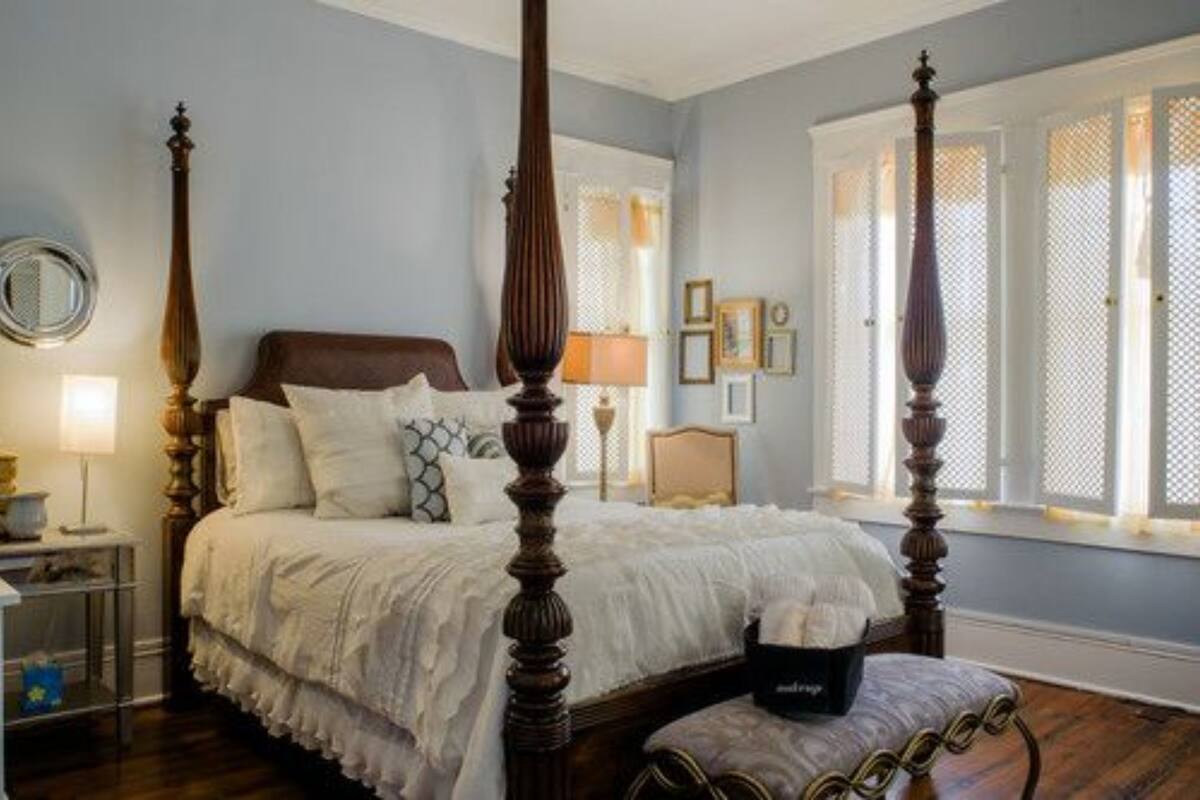 A four-poster bed in a bedroom with a large window and a nightstand with a lamp.