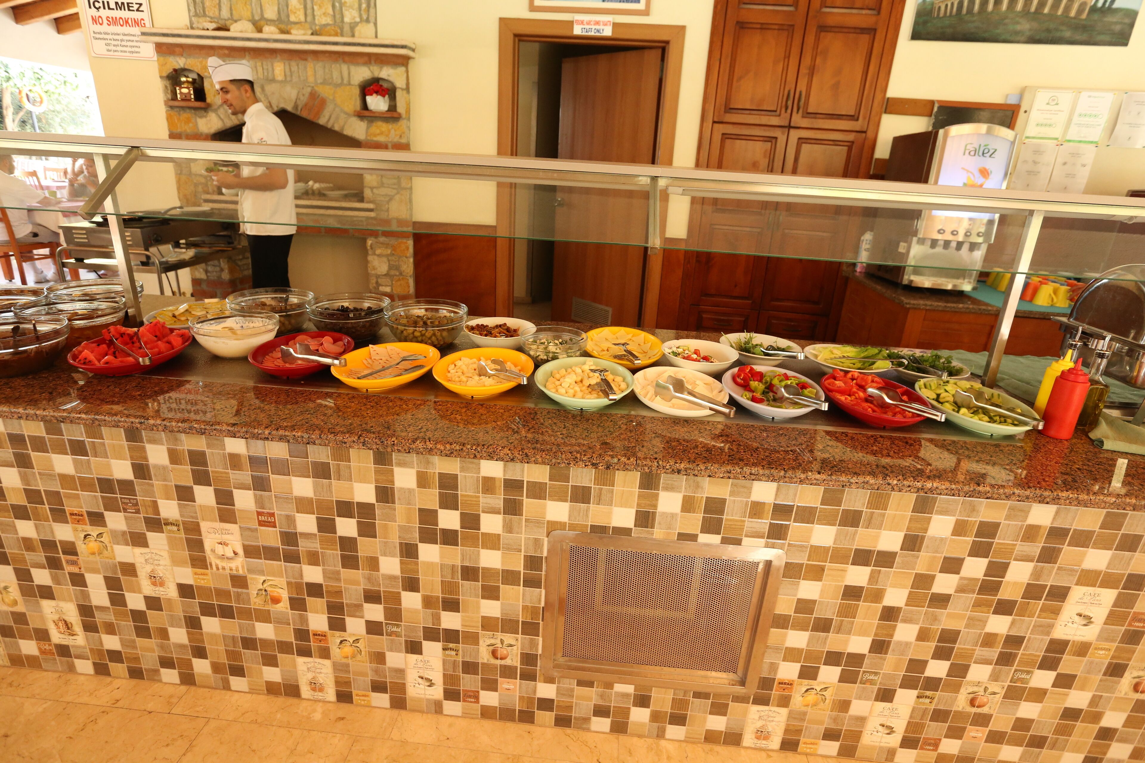 A buffet area with various food dishes displayed in bowls and plates, a chef preparing food, and a tiled backsplash with fruit designs.