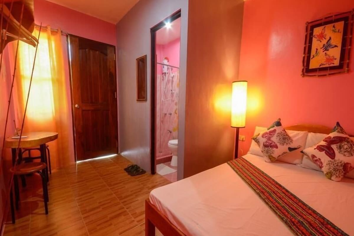 A hotel room with a wooden floor, a bed with floral pillows, a bedside lamp, a wooden door, and a mirror.