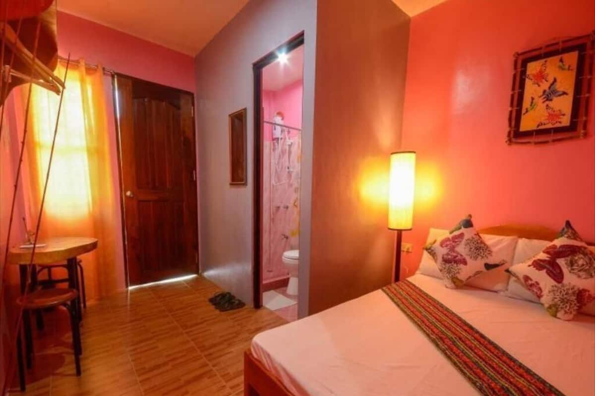 Ein Zimmer mit Holzboden, einem Bett mit Blumenkissen, einer Nachttischlampe und einer Holztür.