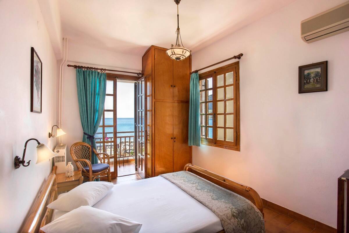A bedroom with a bed, a wooden wardrobe, a chair, a balcony with a view of the sea, and a window with curtains.