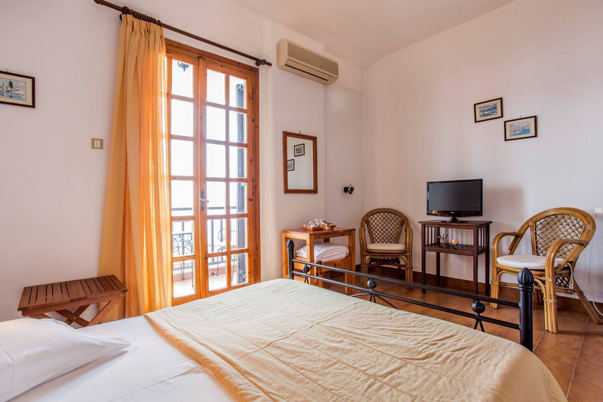 A hotel room with a bed, a television, two wicker chairs, a small table, and a window with curtains.