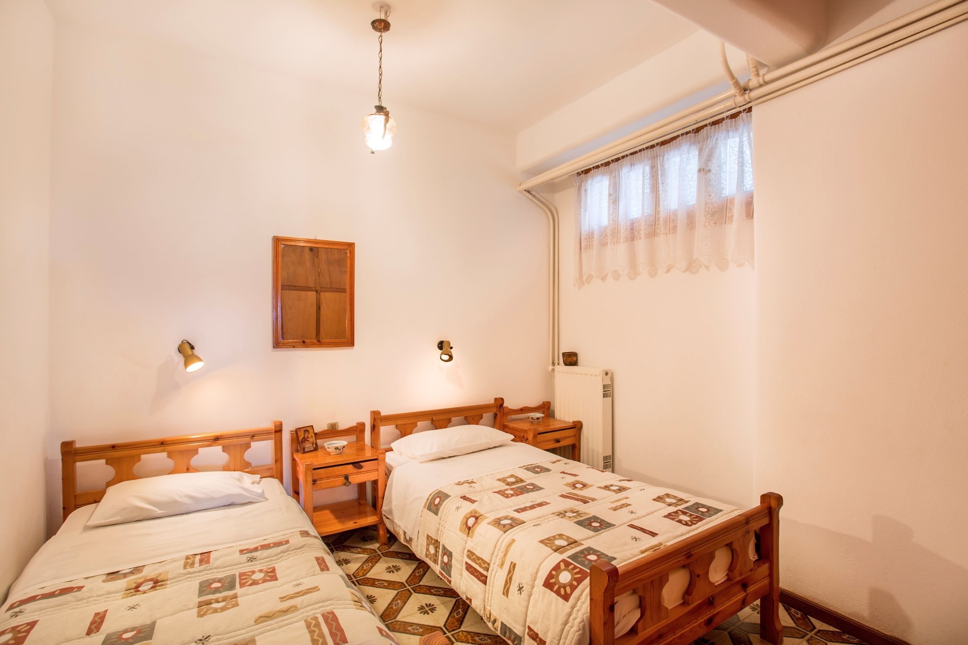 Quarto com duas camas de madeira, uma mesinha de cabeceira de madeira, uma luminária na parede, uma janela com cortinas de renda e um radiador.