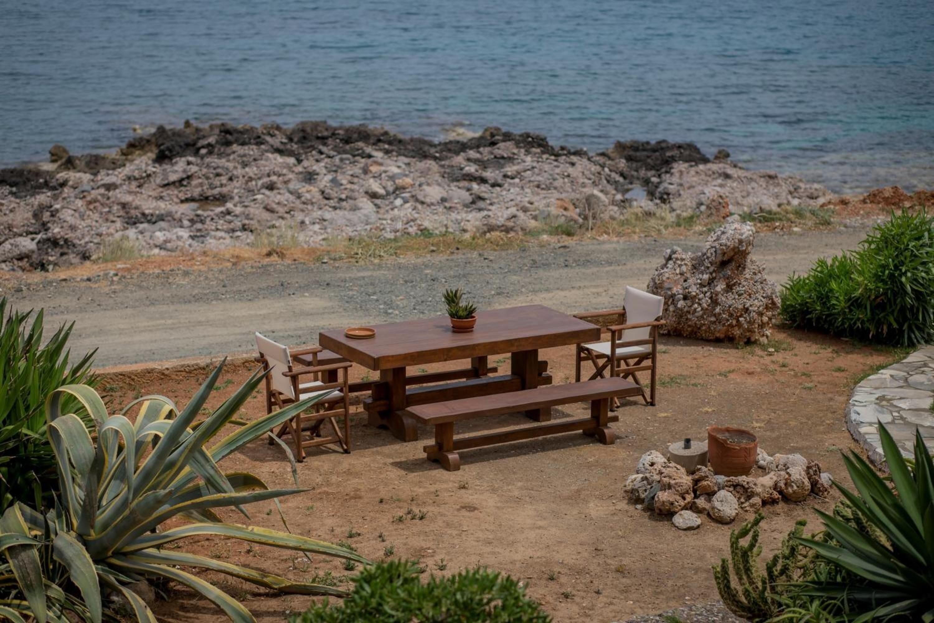 A wooden picnic table with benches and chairs, surrounded by plants and rocks, near a rocky shoreline.