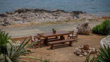 A wooden picnic table with benches and chairs, surrounded by plants and rocks, near a rocky shoreline.