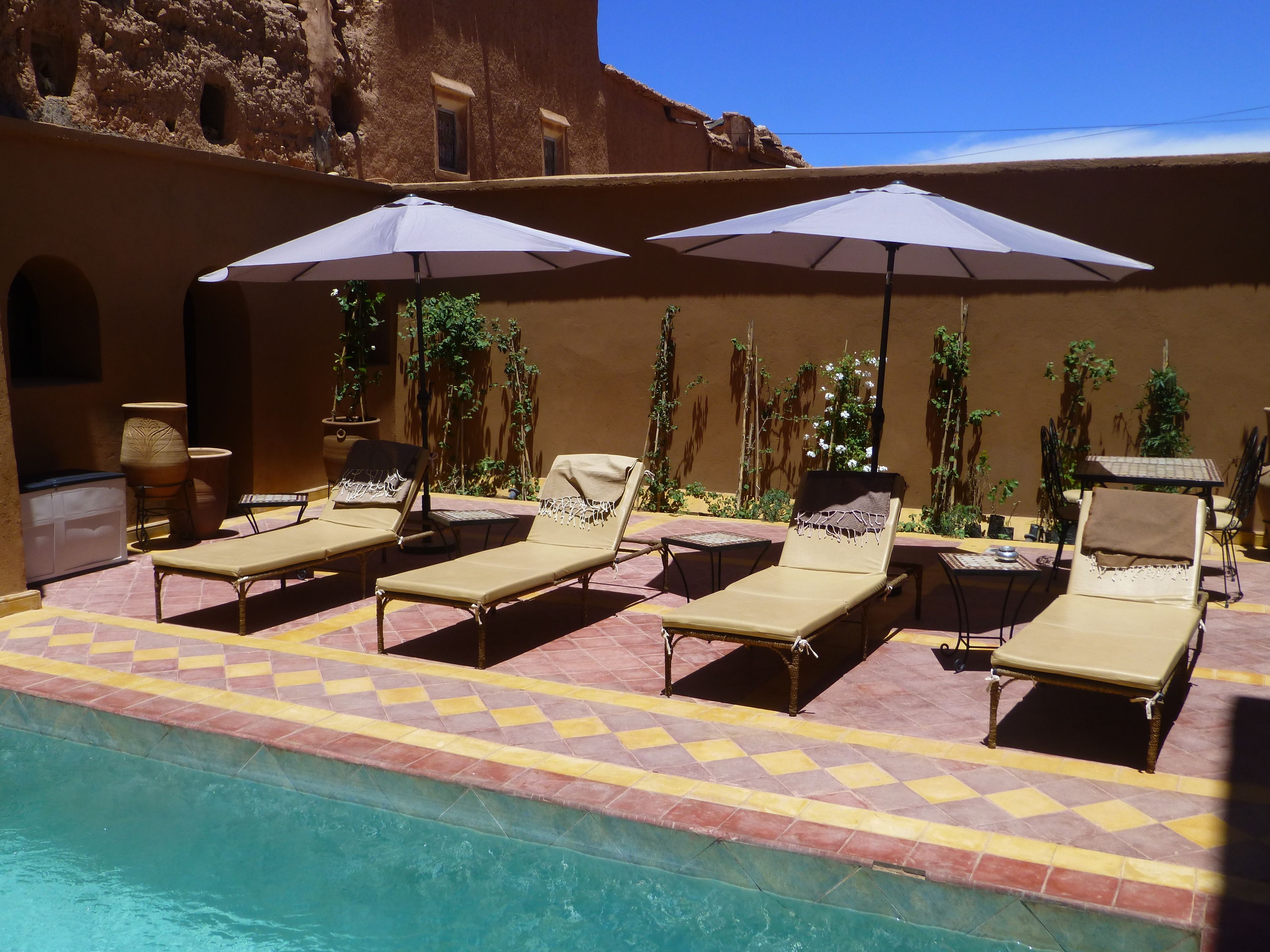 Un área junto a la piscina con sillones de descanso y sombrillas, suelo de baldosas y una piscina.