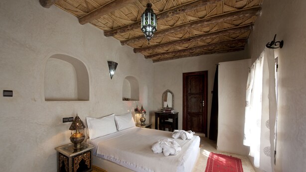 A bedroom with a large bed, a nightstand with a lamp, a red rug, and a wooden ceiling. - Kasbah La Cigogne (Aït Benhaddou)