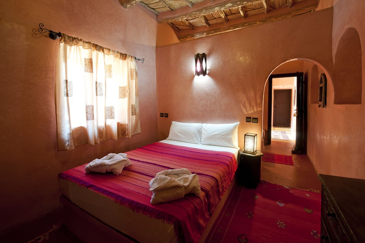 A bedroom with a bed, a window with curtains, a wall-mounted light, and a door leading to another room.