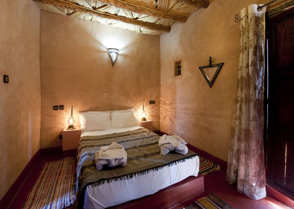 A bedroom with a bed, two bedside lamps, a decorative wall piece, and a window with curtains. - Kasbah La Cigogne (Aït Benhaddou)
