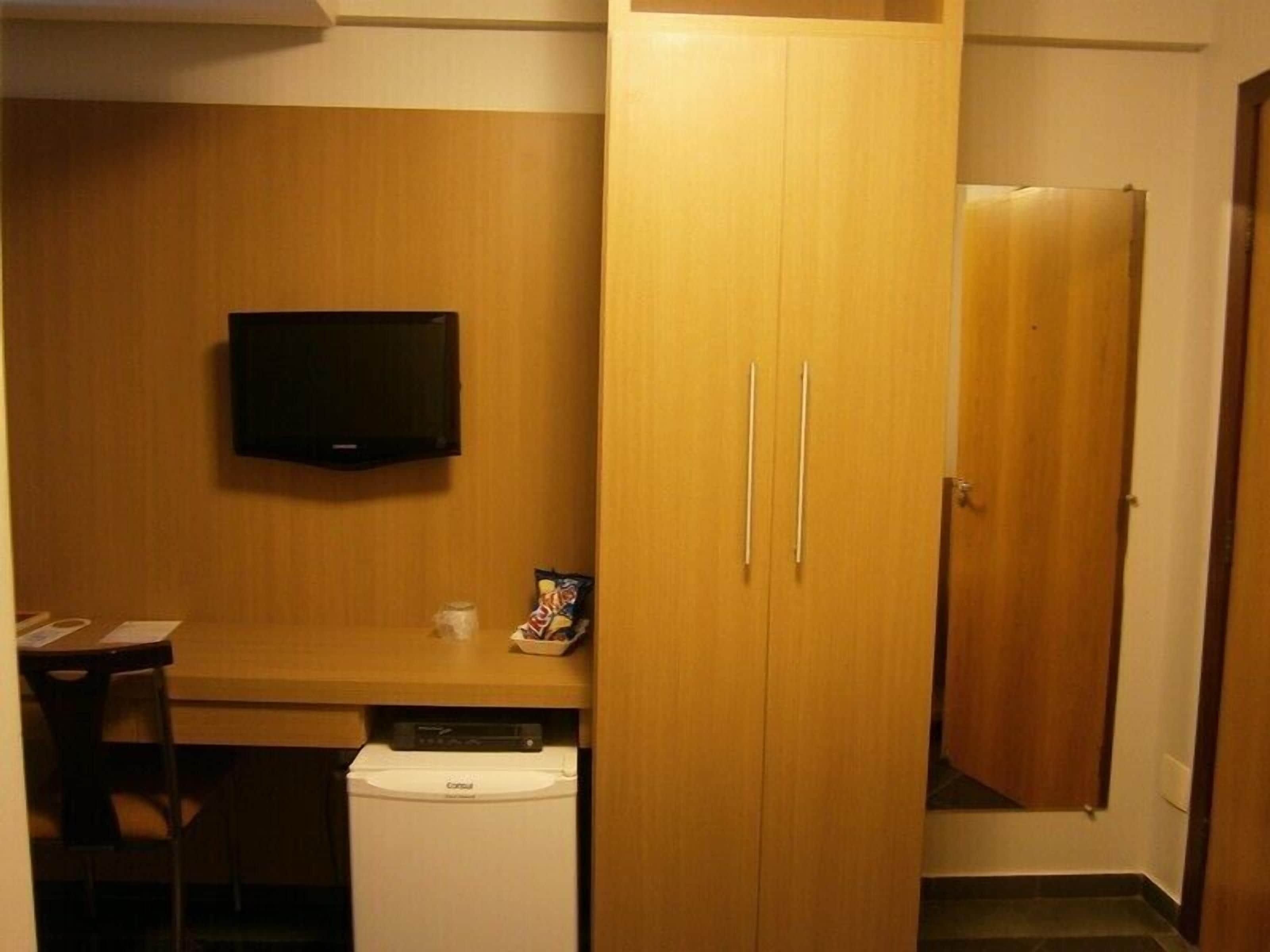 A compact room with a wooden wardrobe, a small table with a chair, a television mounted on the wall, and a washing machine.