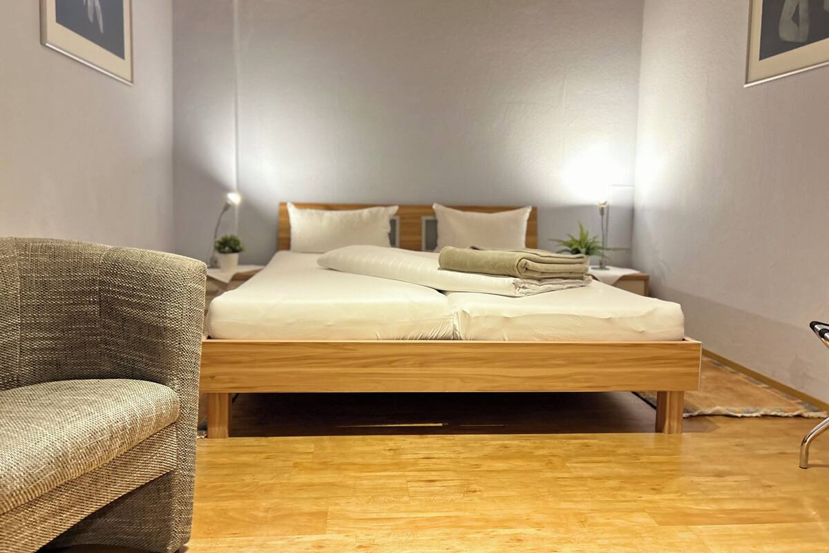 Ein Schlafzimmer mit einem hölzernen Bett, einem Sessel, einem Nachttisch mit Lampe und einer kleinen Pflanze.