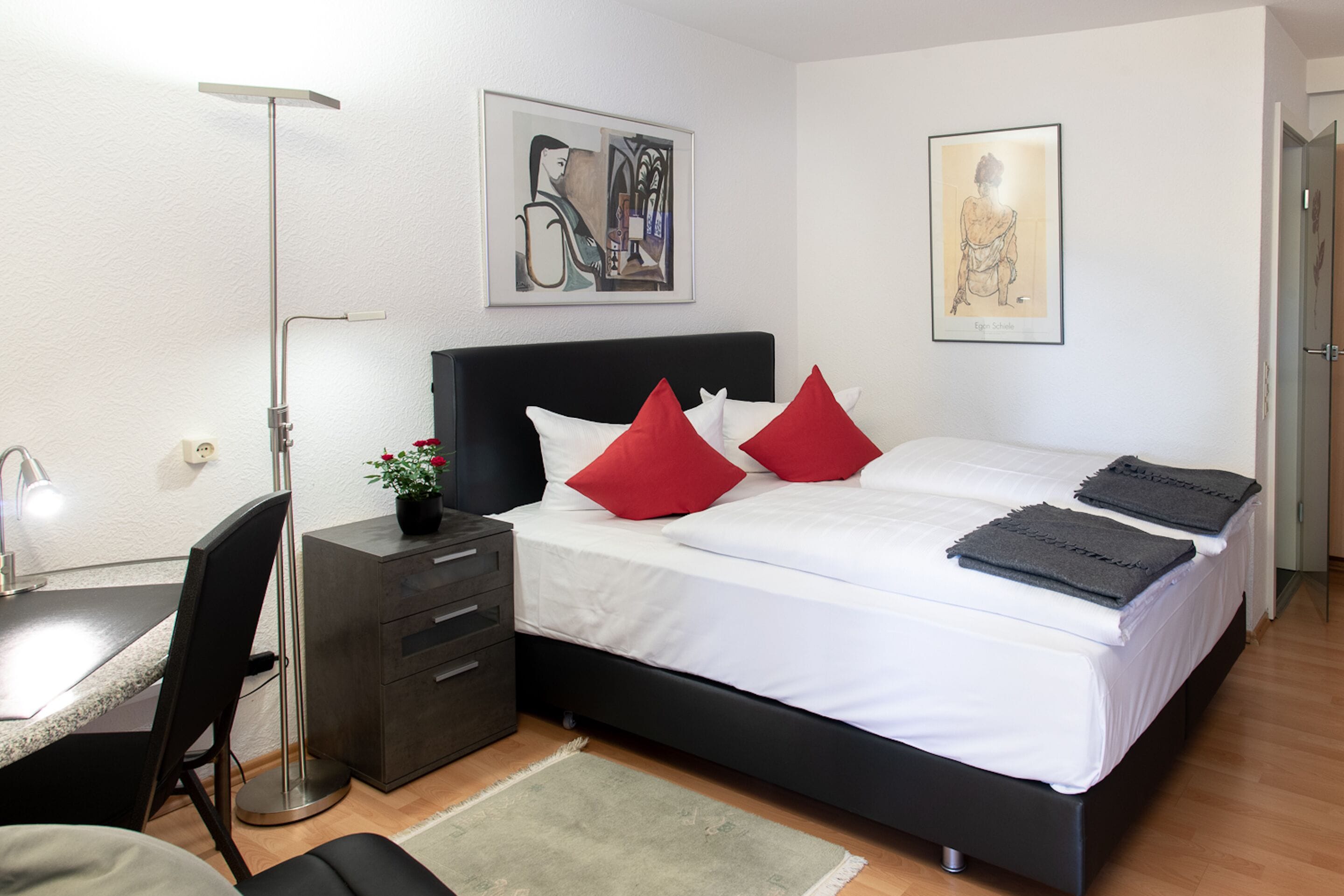 A modern bedroom with a black headboard, white bedding, red pillows, a desk with a chair, a floor lamp, and artwork on the walls.
