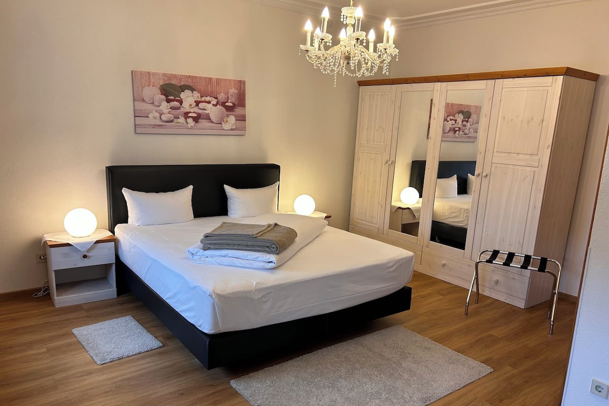 Ein ordentlich bezogenes Bett mit einem gefalteten Handtuch, ein Nachttisch mit einer Lampe, eine Chandelier, ein Kleiderschrank und ein Spiegel.