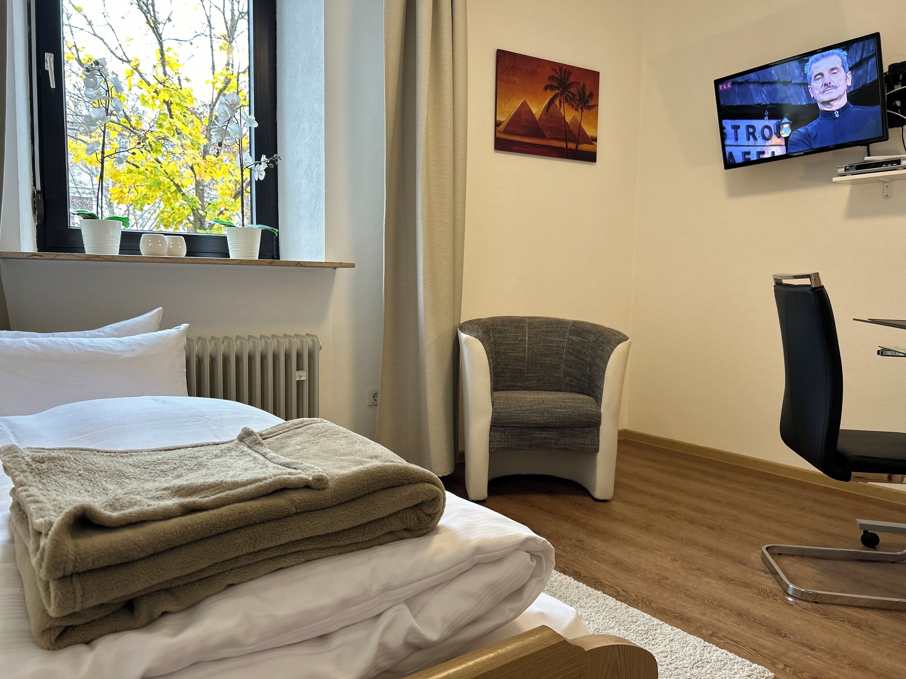 Habitación de hotel con cama, una silla, televisor y ventana con vista a árboles.
