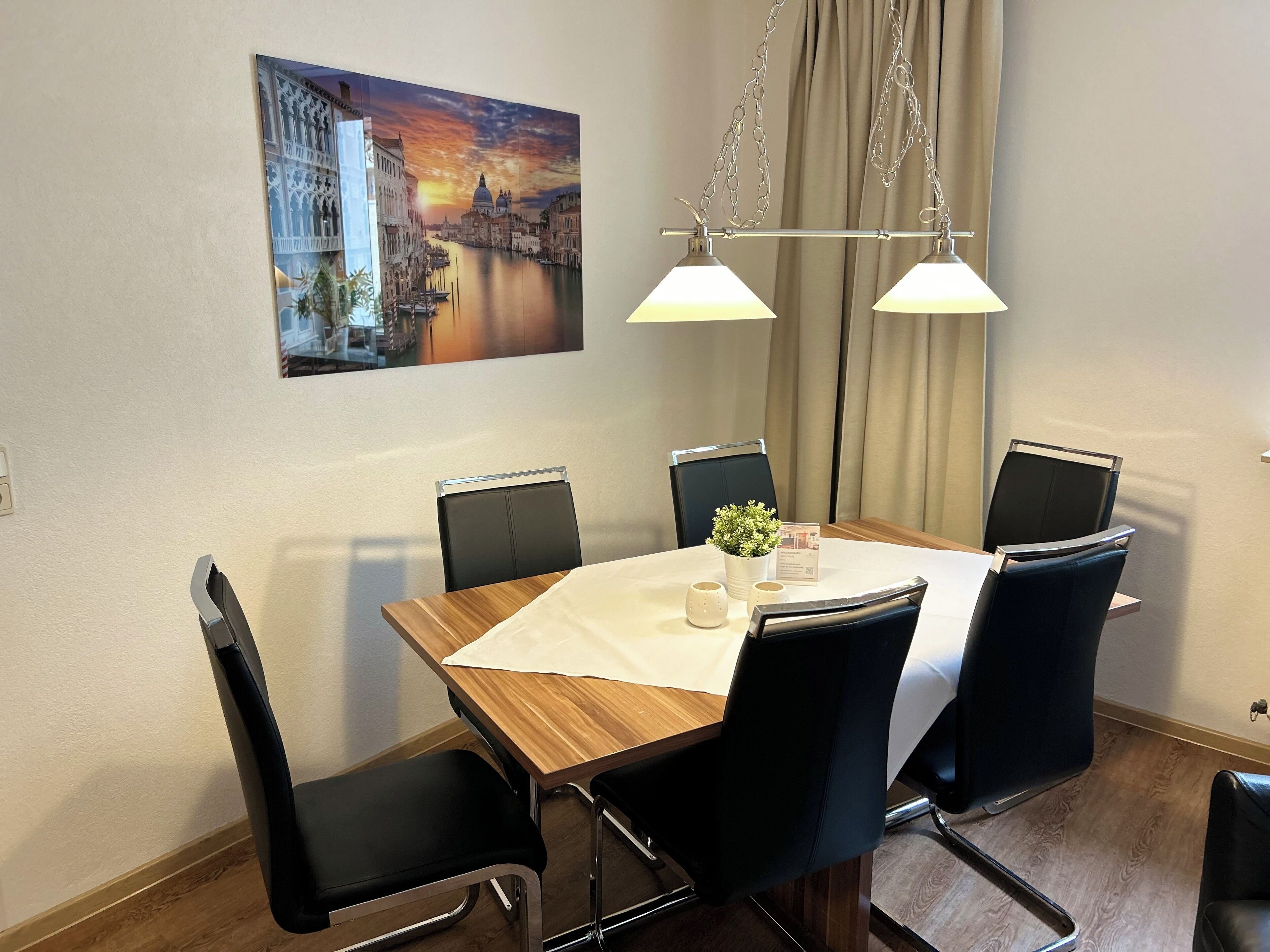 A dining area with a wooden table, black chairs, a white tablecloth, a small potted plant, and a painting of a cityscape on the wall.