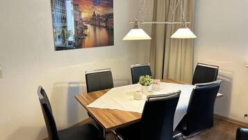 A dining area with a wooden table, black chairs, a white tablecloth, a small potted plant, and a painting of a cityscape on the wall.