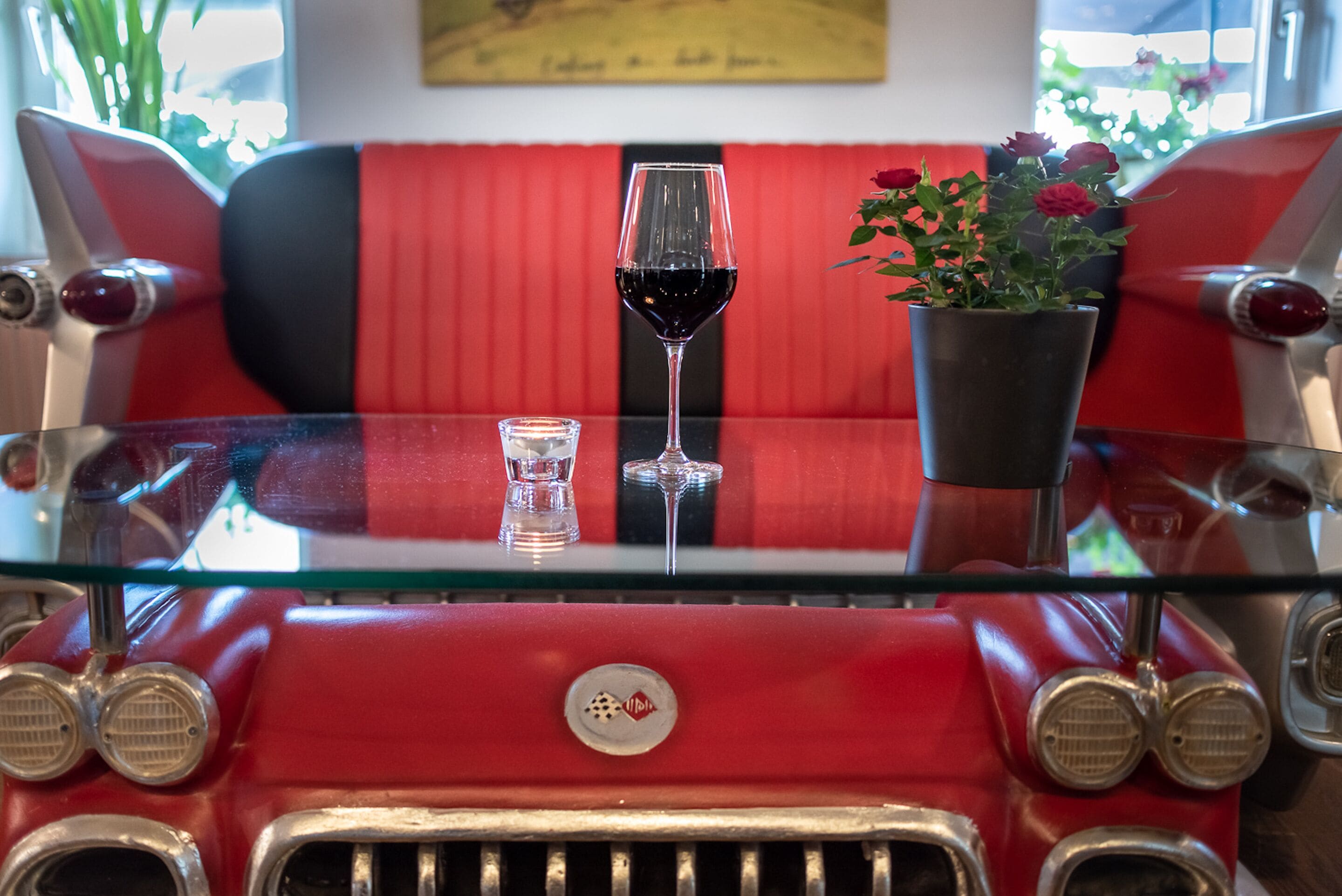 A vintage red car with a glass table in front, a wine glass, and a potted plant with flowers.